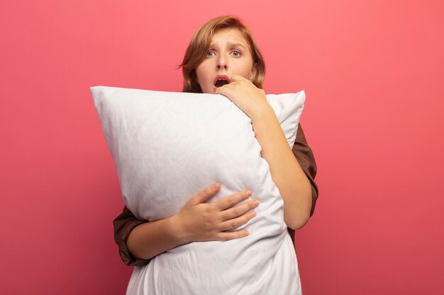 コピースペースとピンクの壁に分離された正面を見て枕を抱き締める心配している若いブロンドの女性