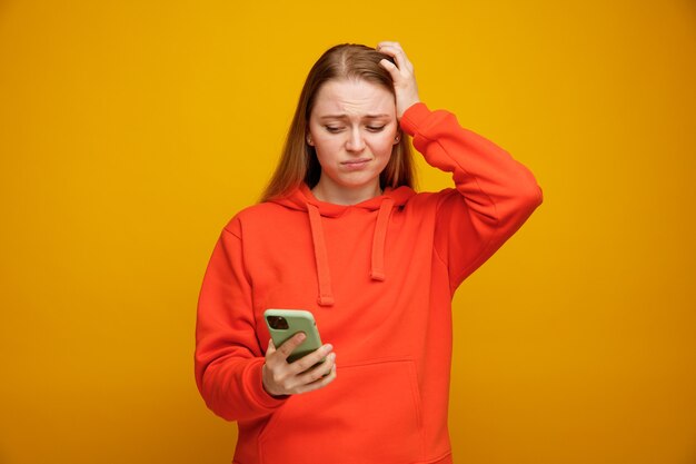 Concerned young blonde woman keeping hand on head holding and looking at mobile phone 