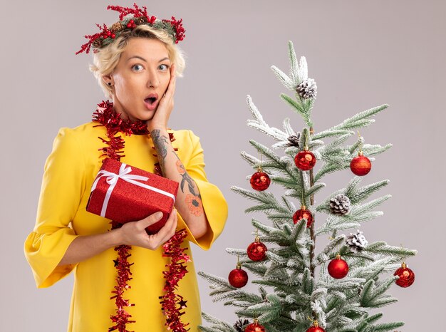 concerned young blonde woman wearing christmas head wreath and tinsel garland around neck standing near decorated christmas tree looking  holding gift package keeping hand on face isolated on white wall