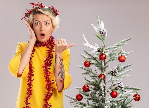 concerned young blonde woman wearing christmas head wreath and tinsel garland around neck standing near decorated christmas tree looking  keeping hand near face pointing at side isolated on white wall