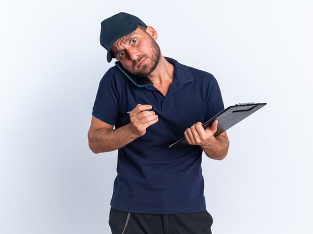 Concerned young caucasian delivery man in blue uniform and cap holding pen and clipboard talking on phone 