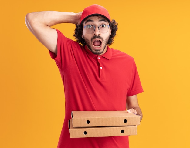 Concerned young caucasian delivery man in red uniform and cap wearing glasses holding pizza packages keeping hand on head isolated on orange wall with copy space