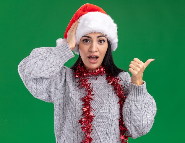 無料写真 クリスマスの帽子と見掛け倒しの花輪を首に身に着けている心配している若い白人の女の子は、緑の背景で隔離された側を指している頭に手を置いてカメラを見て