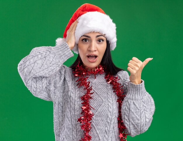 Concerned young caucasian girl wearing christmas hat and tinsel garland around neck  keeping hand on head pointing at side isolated on green wall