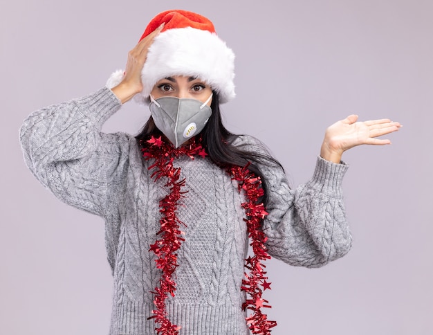 Concerned young caucasian girl wearing christmas hat and tinsel garland around neck with protective mask looking at camera showing empty hand keeping another hand on head isolated on white background