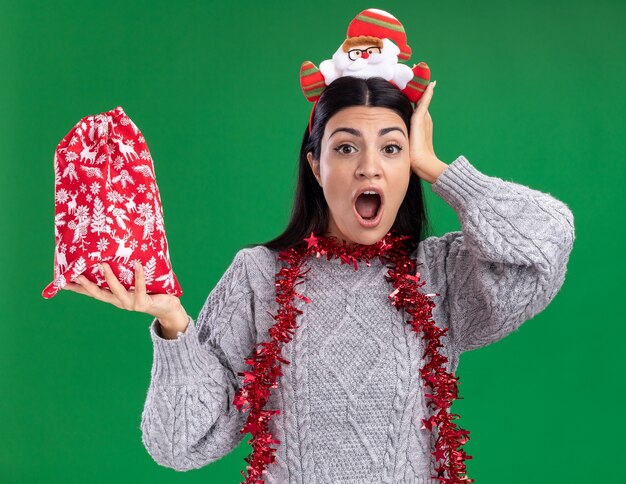 Concerned young caucasian girl wearing santa claus headband and tinsel garland around neck holding christmas gift sack looking at camera keeping hand on head isolated on green background