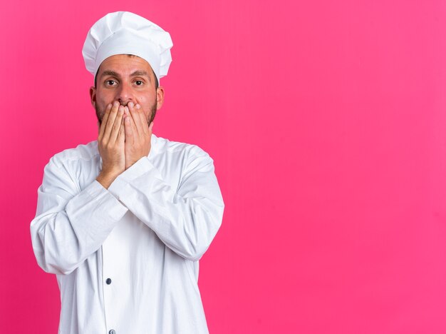 Concerned young caucasian male cook in chef uniform and cap looking at camera covering mouth with hands isolated on pink wall with copy space