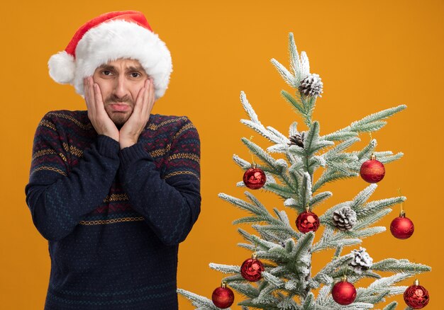 Обеспокоенный молодой кавказец в новогодней шапке стоит возле украшенной елки, держа руки на лице, глядя в камеру, изолированную на оранжевом фоне