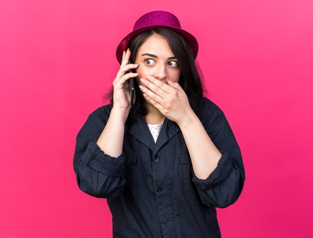 Concerned young caucasian party girl wearing party hat talking on phone looking at side keeping hand on mouth isolated on pink wall