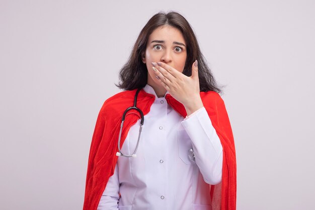 Concerned young caucasian superhero girl in red cape wearing doctor uniform and stethoscope putting hand on mouth 
