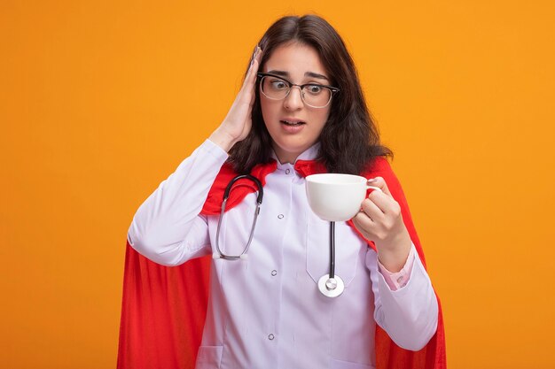 Concerned young caucasian superhero girl wearing doctor uniform and stethoscope with glasses holding and looking at cup of tea keeping hand on head 