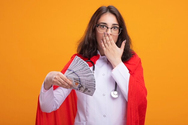 Concerned young caucasian superhero girl wearing doctor uniform and stethoscope with glasses holding money putting hand on mouth 