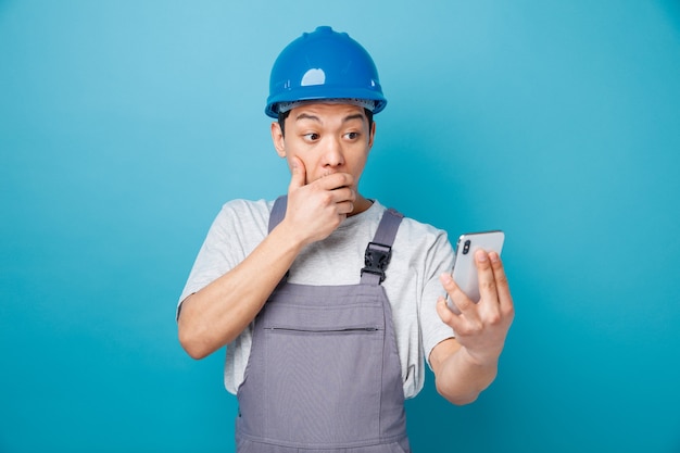 Concerned young construction worker wearing safety helmet and uniform holding and looking at mobile phone keeping hand on mouth 