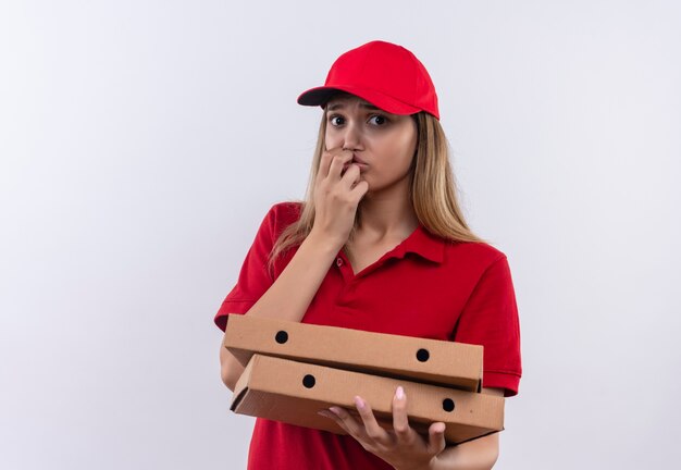 Giovane ragazza preoccupata delle consegne che indossa l'uniforme rossa e il cappuccio che tiene le scatole della pizza e mette la mano sulla bocca isolata sulla parete bianca