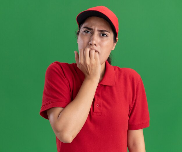 Обеспокоенная молодая доставщица в униформе и кепке грызет ногти на зеленой стене