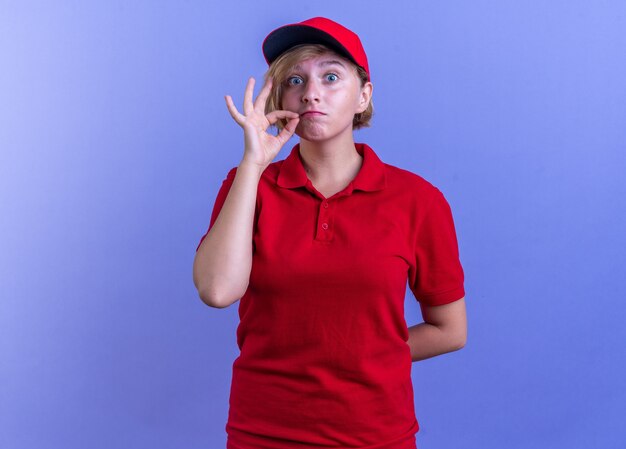 concerned young delivery girl wearing uniform and cap showing silence gesture isolated on blue wall