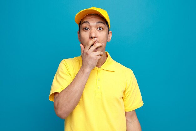 Concerned young delivery man wearing uniform and cap doing oops gesture 