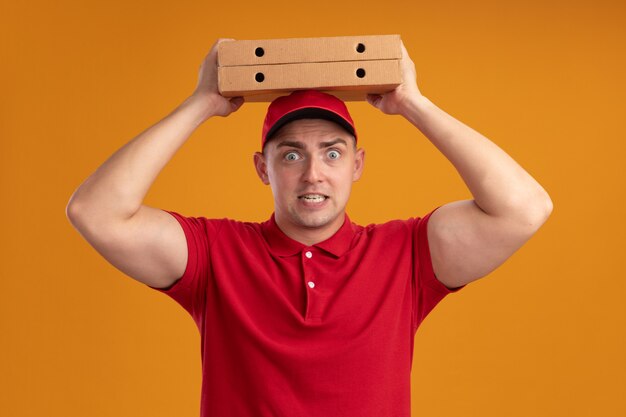 Concerned young delivery man wearing uniform with cap holding pizza boxes on head isolated on orange wall