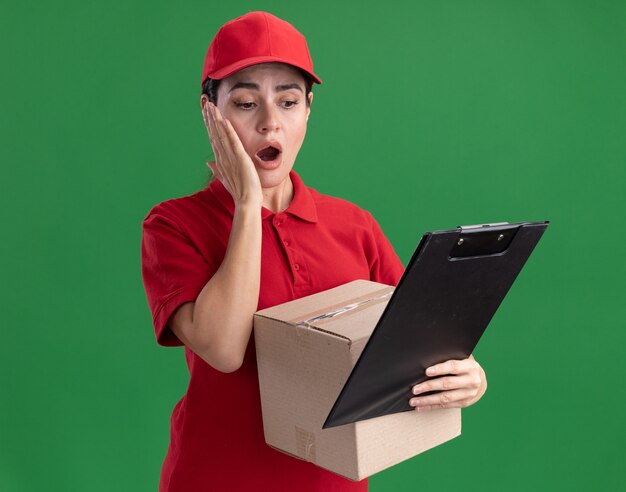 Обеспокоенная молодая женщина-доставщик в униформе и кепке держит картонную коробку и буфер обмена, глядя в буфер обмена