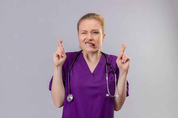 Обеспокоенный молодой врач в фиолетовом медицинском халате и стетоскопе скрещивает пальцы на изолированной белой стене