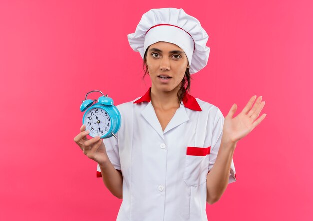 Concerned young female cook wearing chef uniform holding alarm clock spread hand on isolated pink wall with copy space