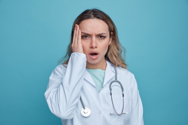 Concerned young female doctor wearing medical robe and stethoscope around neck keeping hand on face 