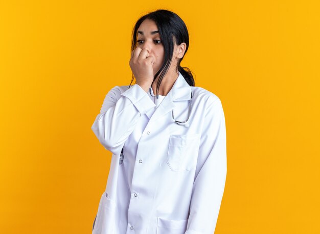 Обеспокоенная молодая женщина-врач в медицинском халате со стетоскопом, положив руку на подбородок, изолированную на желтой стене