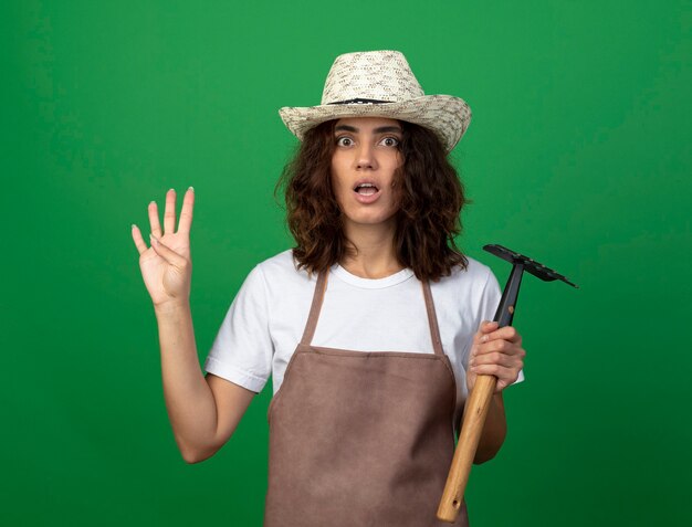 Concerned young female gardener in uniform wearing gardening hat holding rake showing four