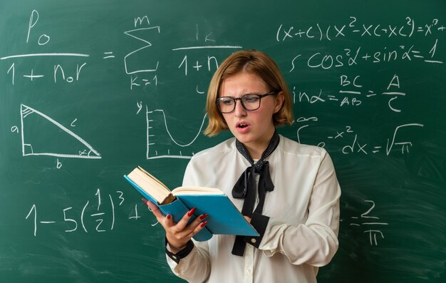 Concerned young female teacher wearing glasses standing in front blackboard reading book in classroom