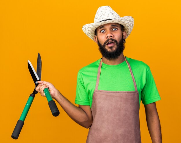 Concerned young gardener afro-american guy wearing gardening hat holding clippers isolated on orange wall