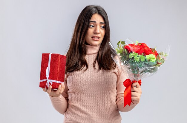 Concerned young girl on valentines day holding gift box looking at bouquet in her hand isolated on white background