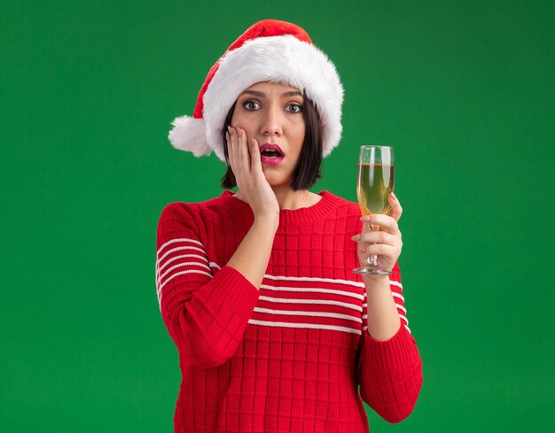 Concerned young girl wearing santa hat holding glass of champagne looking at camera keeping hand on face isolated on green background with copy space