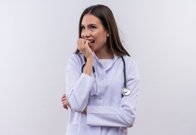Concerned young girl wearing stethoscope medical gown put his hand on chin on isolated white wall