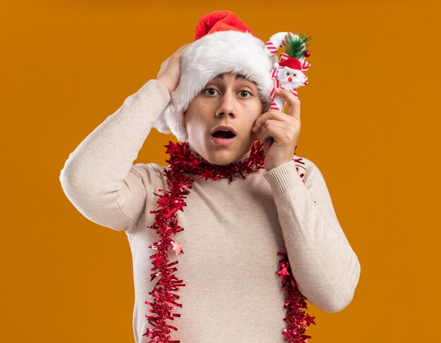 心配している若い男は、黄色の背景で隔離の頭に手を置くクリスマスキャンディーを保持している首に花輪とクリスマス帽子をかぶっています