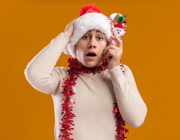 Concerned young guy wearing christmas hat with garland on neck holding christmas candy putting hand on head isolated on yellow background