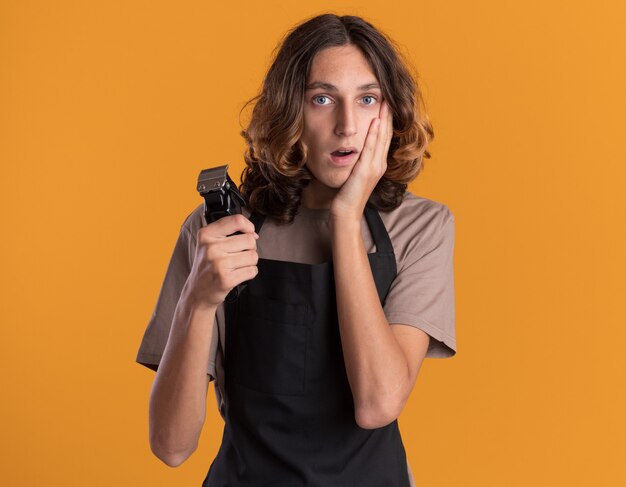 Concerned young handsome barber wearing uniform holding hair clippers keeping hand on face