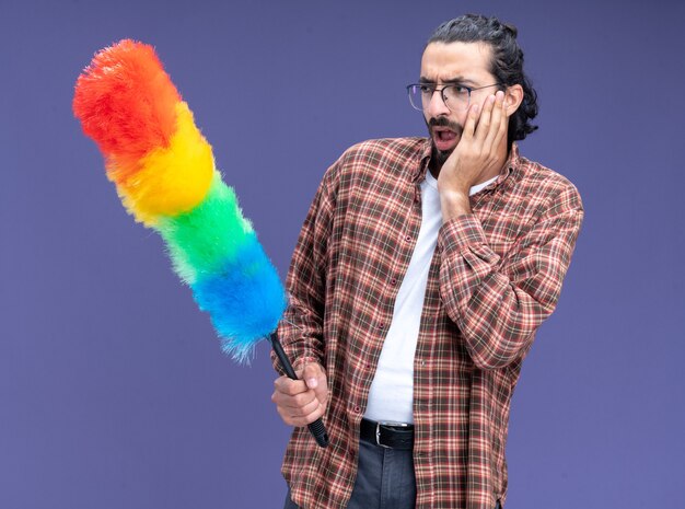 Concerned young handsome cleaning guy wearing t-shirt holding and looking at feather duster putting hand on cheek isolated on blue wall