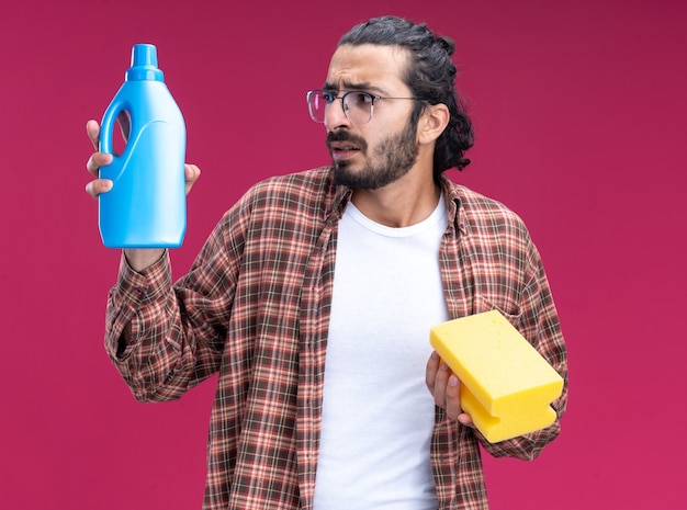 Concerned young handsome cleaning guy wearing t-shirt holding sponge and looking at cleaning agent in his hand isolated on pink wall