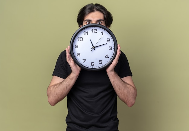 Concerned young handsome guy wearing black t-shirt covered face with wall clock isolated on olive green wall