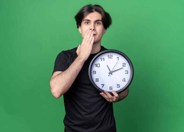 Обеспокоенный молодой красивый парень в черной футболке, держащий настенные часы, прикрыл рот рукой, изолированной на зеленой стене с копией пространства