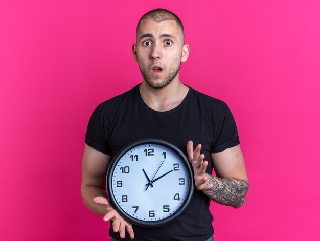 ピンクの背景で隔離の壁時計を保持している黒いTシャツを着ている心配している若いハンサムな男