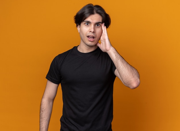 Concerned young handsome guy wearing black t-shirt putting hand on cheek isolated on orange wall with copy space