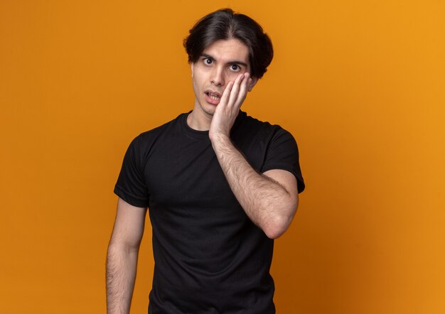 Concerned young handsome guy wearing black t-shirt putting hand on cheek isolated on orange wall