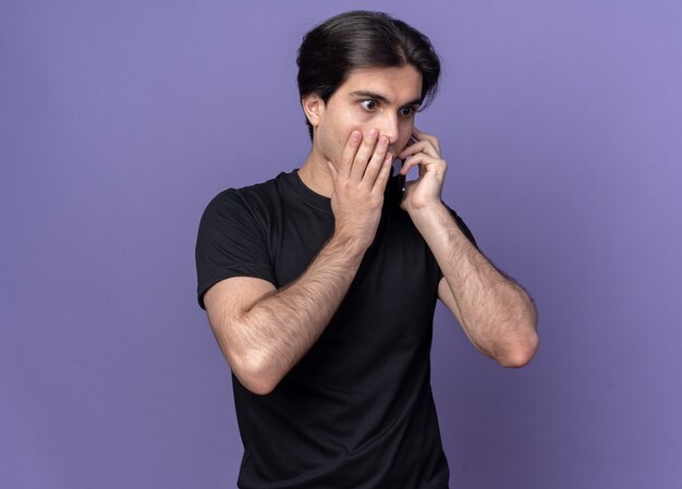 Concerned young handsome guy wearing black t-shirt speaks on phone covered mouth with hand isolated on purple wall