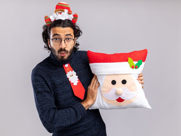 Concerned young handsome guy wearing christmas tie with hair hoop holding christmas pillow isolated on white wall