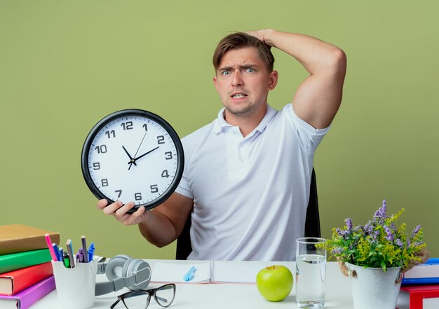 Concerned young handsome male student sitting at desk with school tools holding wall clock and putting hand on head isolated on olive green