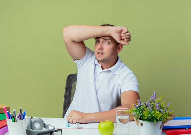 Concerned young handsome male student sitting at desk with school tools putting arm on forehead isolated on olive green