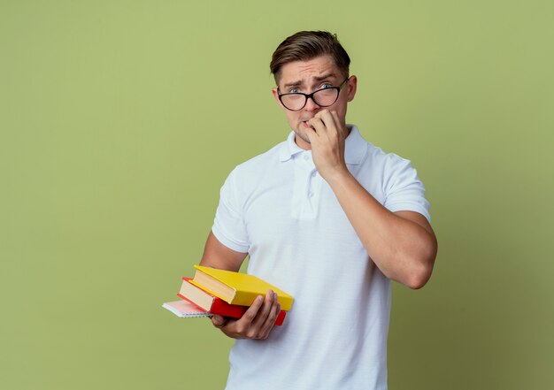 Concerned young handsome male student wearing glasses holding books and bites nails isolated on olive green background