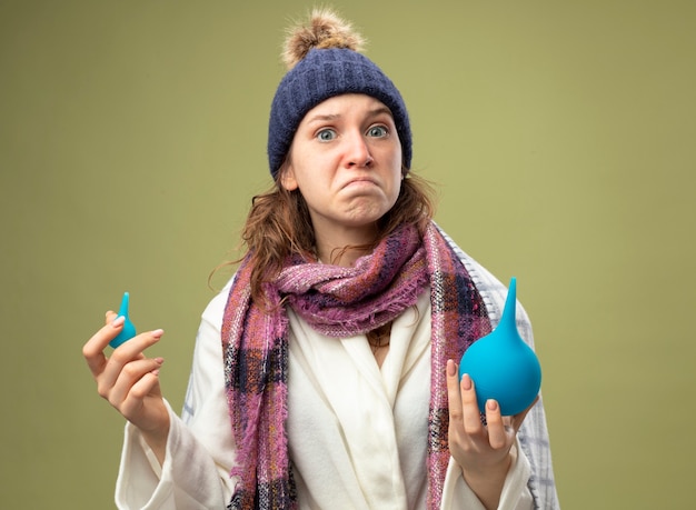 Free photo concerned young ill girl wearing white robe and winter hat with scarf wrapped in plaid holding enemas isolated on olive green