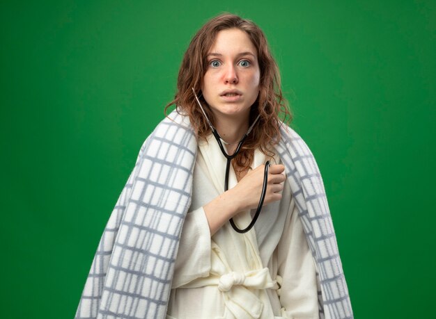 Concerned young ill girl wearing white robe wrapped in plaid listening to her own heartbeat with stethoscope isolated on green